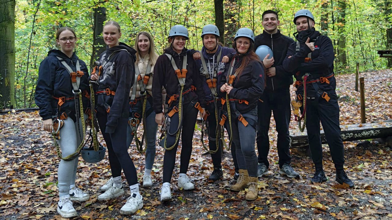 8 junge Menschen stehen in einem Wald. Sie tragen Kletterausrüstung, vier Personen tragen einen Helm.