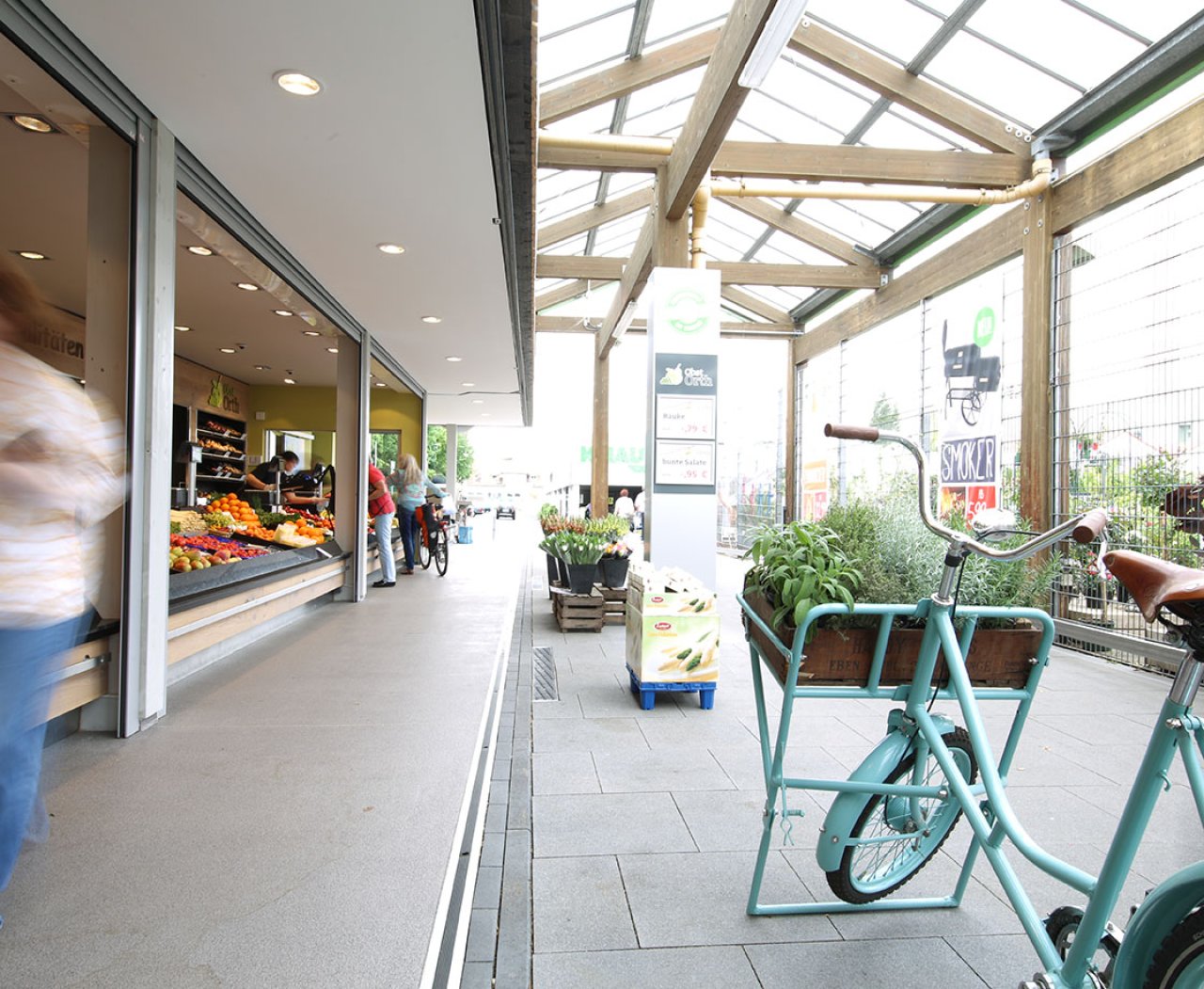 Überdachter Gehweg mit Fahrradständer und grünem Fahrrad neben einem Obst- und Gemüseladen.
