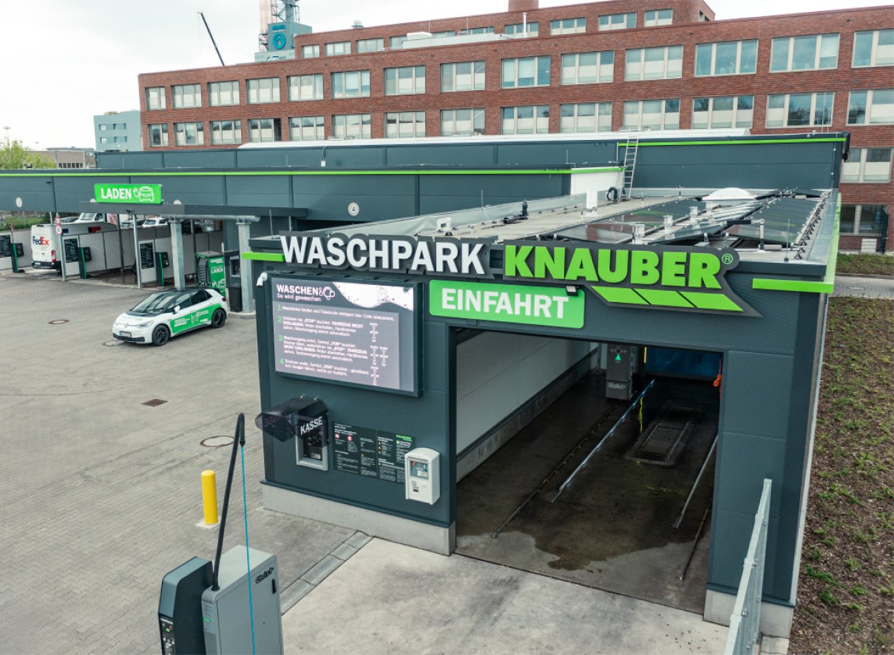 Einfahrt zum Waschpark Knauber mit grüner Beschilderung und einem weißen Auto auf dem Parkplatz vor einem mehrstöckigen Gebäude.