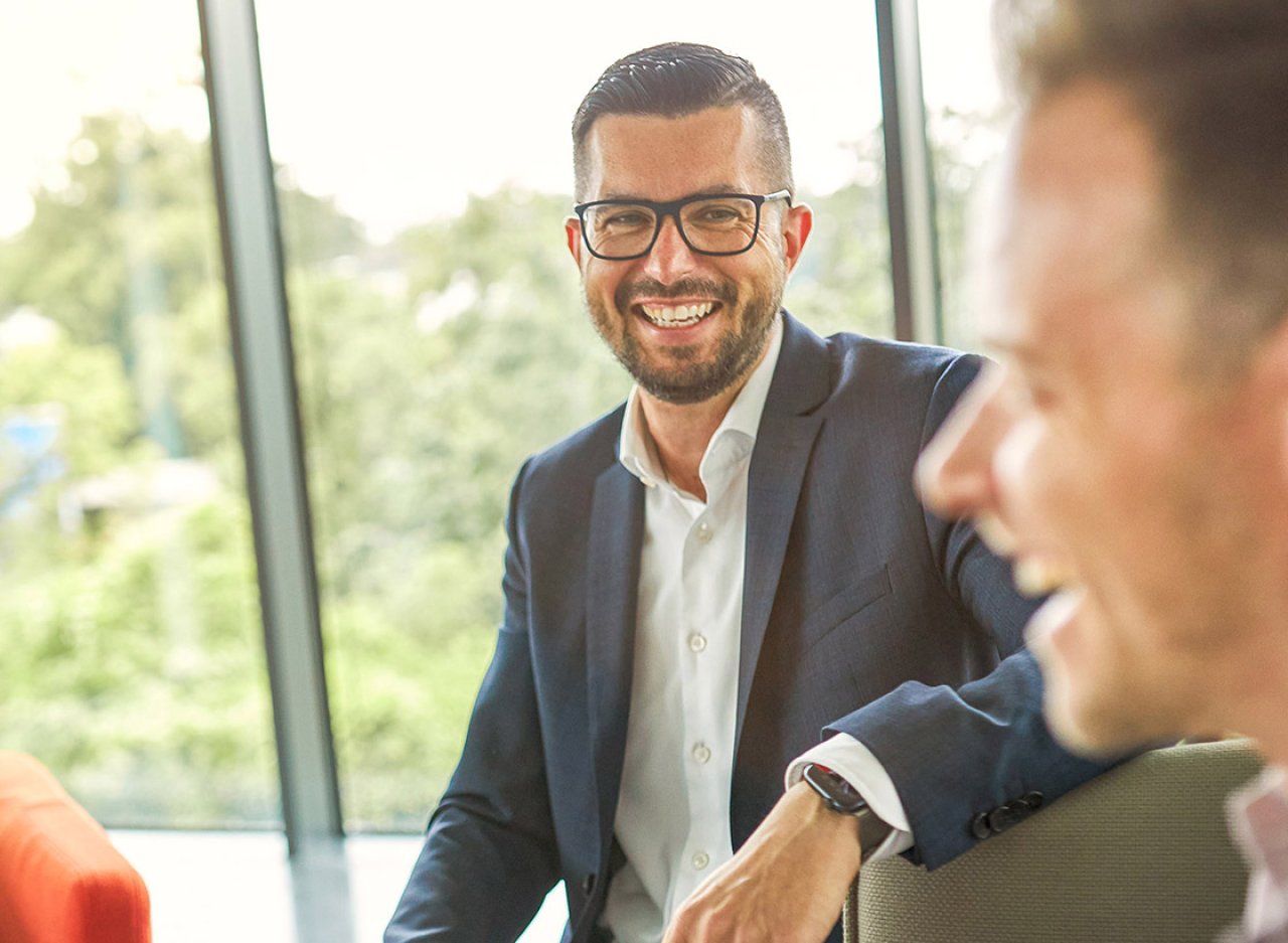 Zwei Männer in Business-Kleidung unterhalten sich und lachen in einem hellen Büro mit großen Fenstern.