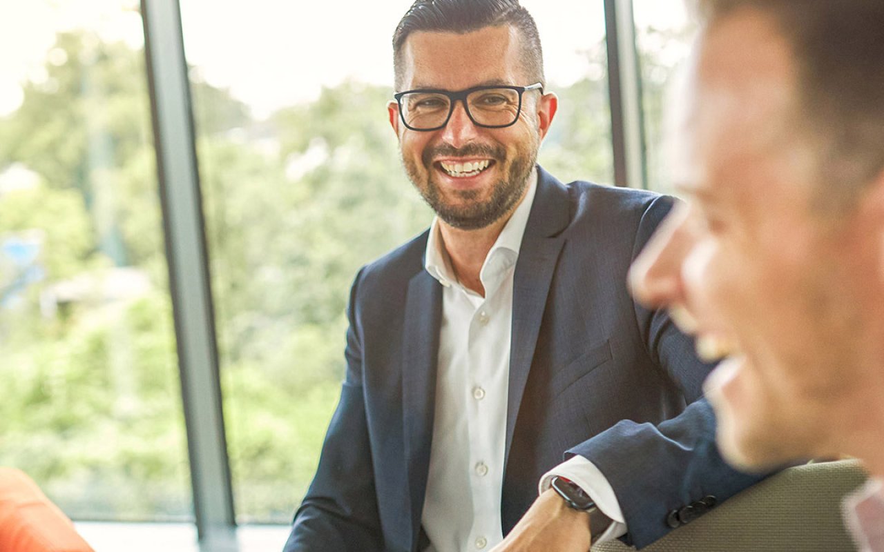 Zwei Männer in Business-Kleidung unterhalten sich und lachen in einem hellen Büro mit großen Fenstern.