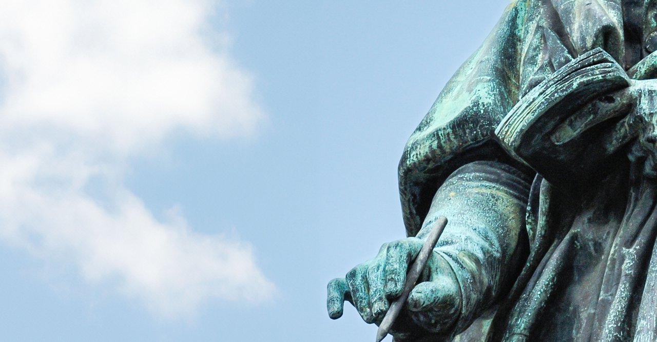 Detailaufnahme einer grünen Bronzestatue mit ausgestreckter Hand vor blauem Himmel mit Wolken.