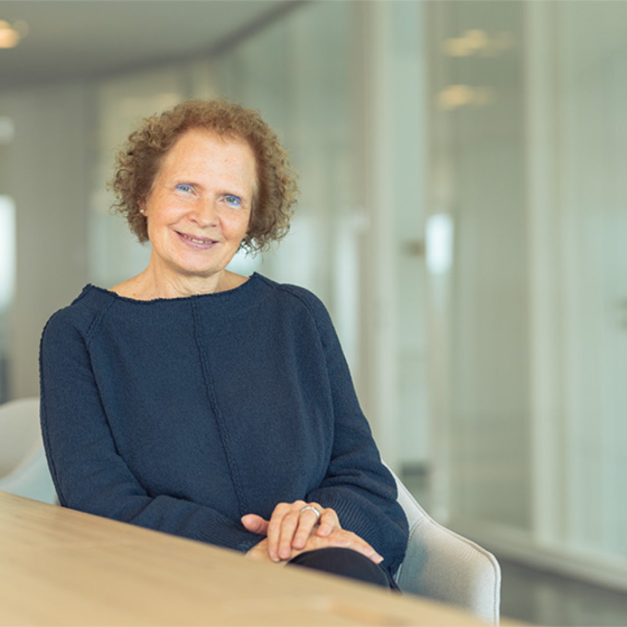 Frau mit lockigem Haar und blauem Pullover sitzt lächelnd an einem Tisch in einem hellen Büro.