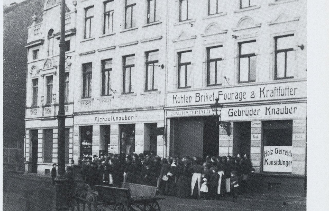 Menschenmenge vor einem Geschäftshaus mit Schildern für Kohlen, Briketts, Futter und Gebrüder Knauber in einer historischen Straßenszene.