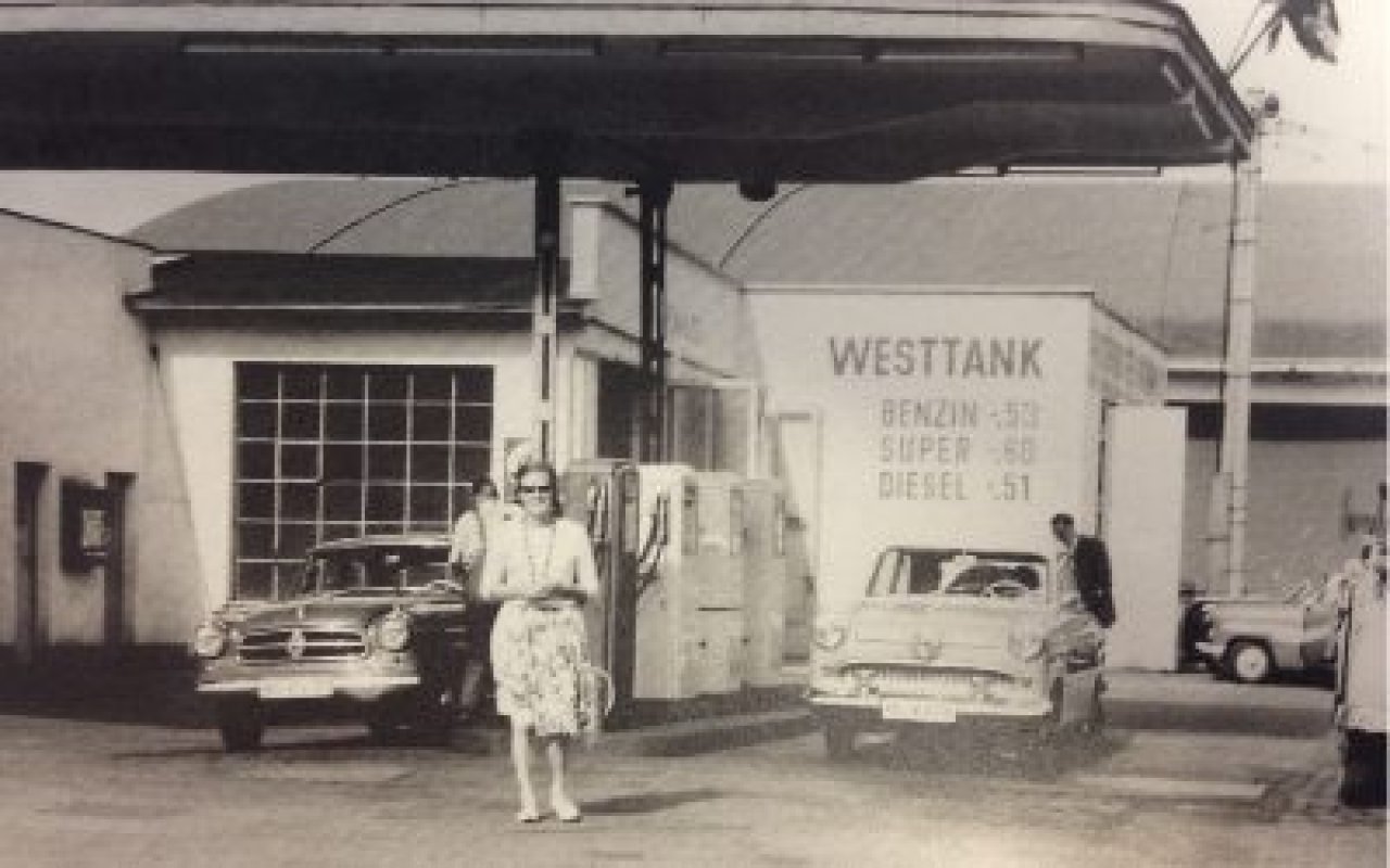 Historisches Bild einer Dame mit Schal vor einer Tankstelle, schwarz-weiß Aufnahme.