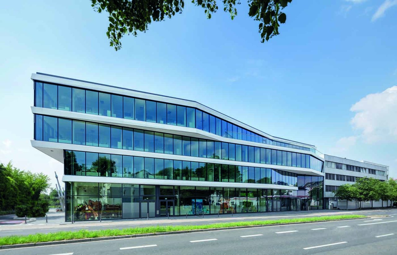 Modernes Bürogebäude mit mehreren Etagen und Glasfassade vor blauem Himmel.