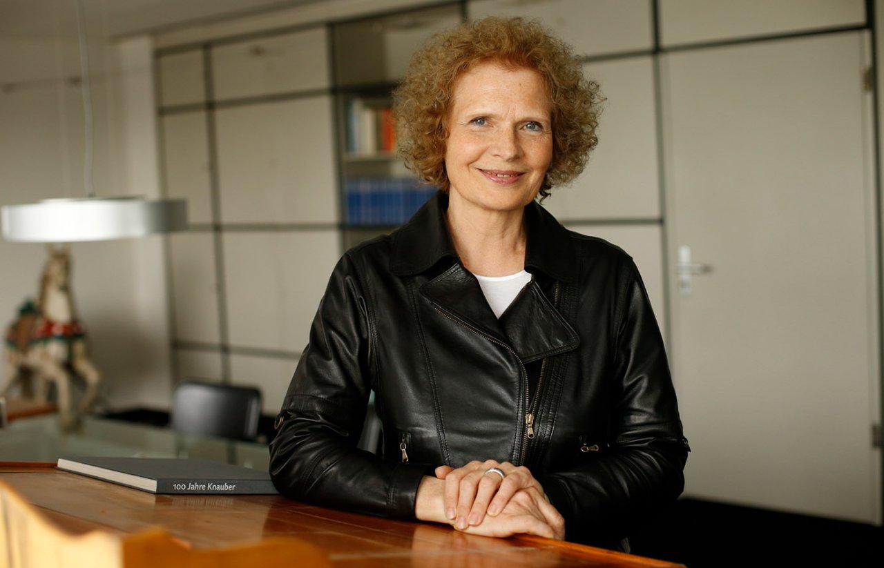 Junge Frau mit blonden Locken und schwarzer Jacke sitzt an einem Holztisch.