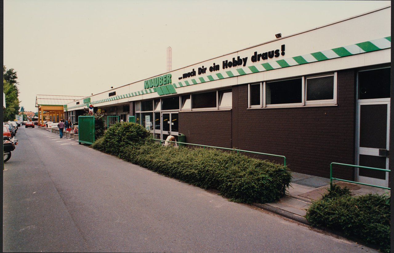 Langes Gebäude mit grün-weißem Streifen und Schriftzug 'MAUSER ... macht dir ein Hobby draus!' an der Fassade, Straße mit parkenden Autos und Büschen davor.