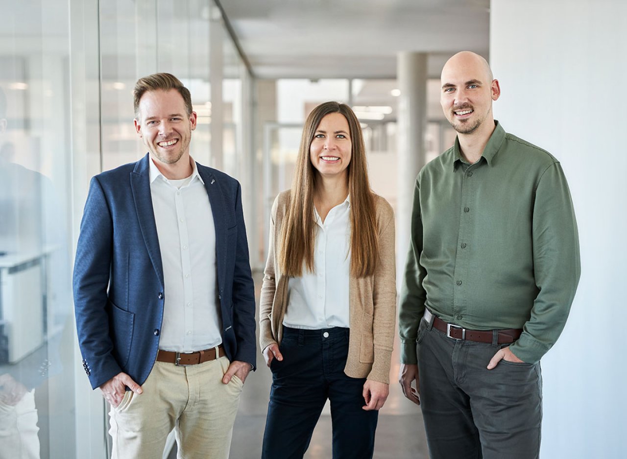 Zwei Männer und eine Frau in Businessoutfits stehen in einem Bürogang und lächeln in die Kamera.