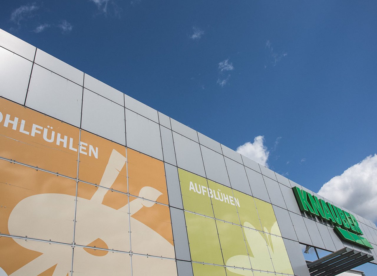 Gebäudefassade mit großen farbigen Plakaten und den Worten 'WOHLFÜHLEN' und 'AUFBLÜHEN' unter blauem Himmel.