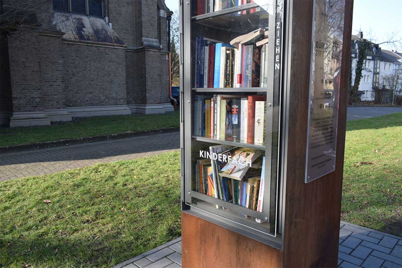 Ein Bücherschrank drußen auf dem Rasen vor einem großen Gebäude.