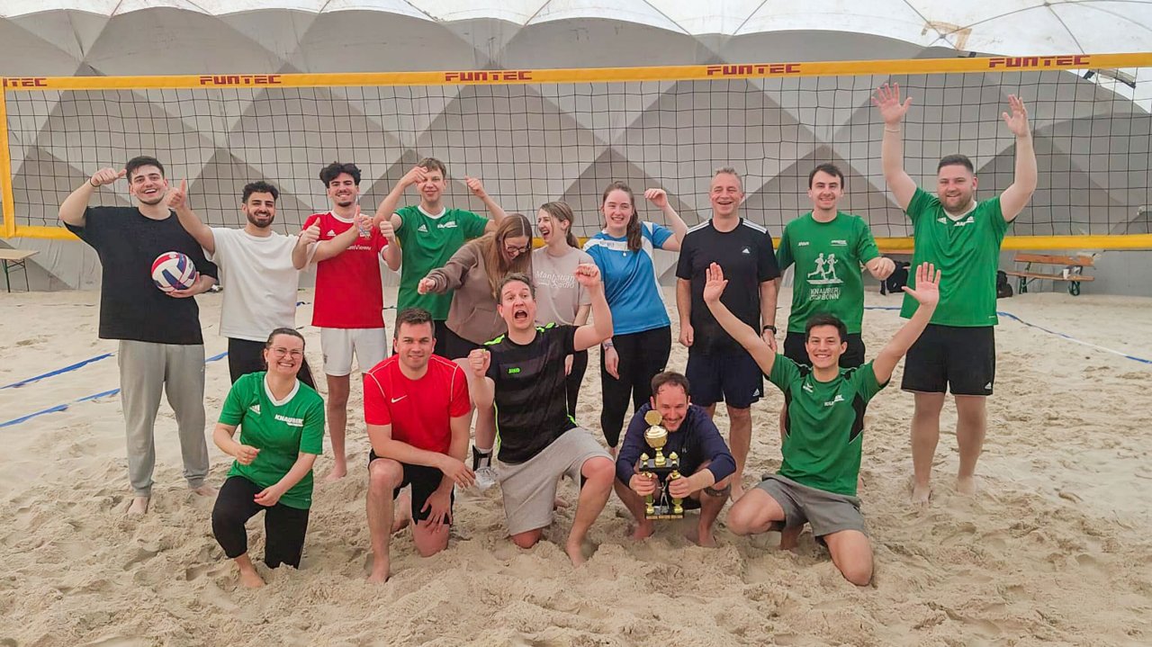 Eine Gruppe junger Menschen in bunten Sportoutfits stehen vor einem Netz auf einem Beachvolleyballfeld.