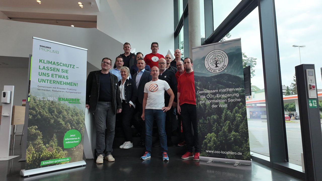 Gruppe von elf Personen vor zwei Bannern mit Umweltschutzbotschaften in einem modernen Gebäude mit großen Fenstern.